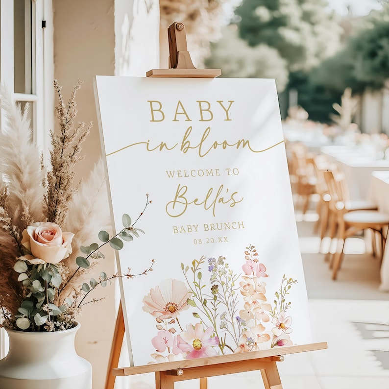 Może przedstawiać: Biały znak na drewnianym sztaludze z napisem "BABY in bloom" złotym skryptem. Znak wita gości na Baby Brunch Belli, z datą poniżej. Kwiatowe ilustracje w kolorze r&oacute;żowym, brzoskwiniowym i fioletowym zdobią d&oacute;ł.