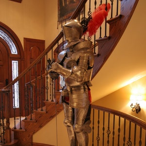 May include: A silver suit of armor with a red and black plume stands on a black pedestal in front of a wooden staircase.