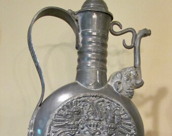 May include: A pewter pitcher with a decorative design featuring a coat of arms and swirling patterns. The pitcher has a tall, slender base and a spout with a handle.