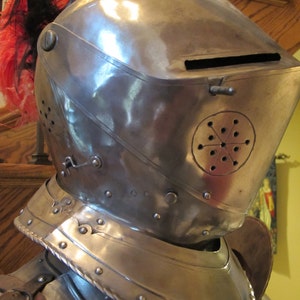 May include: A silver helmet with a plume of red and black feathers. The helmet has a visor and a decorative design on the side.