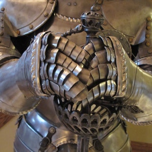 May include: Close-up of a suit of armor with a metal gauntlet on each hand. The gauntlets are detailed with intricate metalwork and are holding a sword.