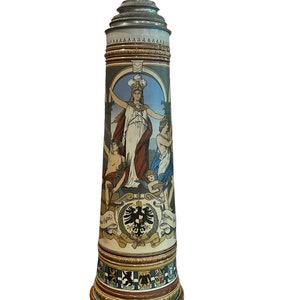 Peut inclure: Une grande chope en céramique avec un design détaillé. La chope représente une femme en robe blanche tenant une couronne de laurier, entourée d'autres personnages. La chope présente également un blason avec un aigle noir et le texte "Einigkeit und Recht und Freiheit".