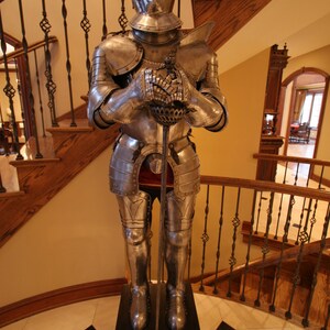 May include: A full suit of silver armor with a red plume on the helmet, holding a sword, standing on a black wooden base.