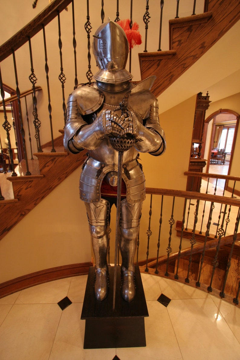 May include: A full suit of silver armor with a red plume on the helmet, holding a sword, standing on a black wooden base.