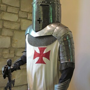 May include: A knight in full armor, wearing a helmet with a visor and a chainmail shirt. The knight is holding a sword and has a red cross on his chest.