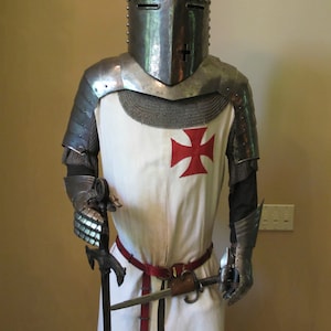 May include: A knight in full armor, wearing a white tunic with a red cross, holds a sword. The knight is wearing a helmet with a visor.