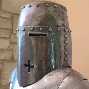 May include: A close-up of a medieval helmet with a visor and a cross on the front. The helmet is made of metal and has a textured surface.