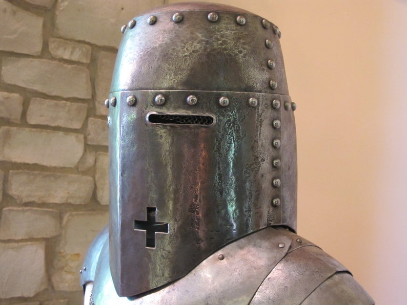 May include: A close-up of a medieval helmet with a visor and a cross on the front. The helmet is made of metal and has a textured surface.