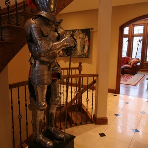 May include: A full-body suit of silver armor with a red plume on the helmet. The armor is standing on a black pedestal in a room with a wooden staircase and a large window.
