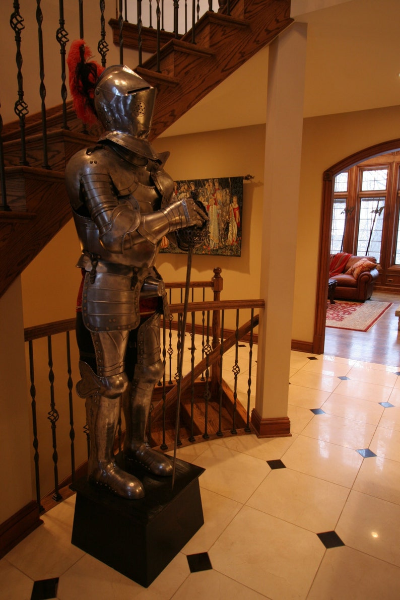 May include: A full-body suit of silver armor with a red plume on the helmet. The armor is standing on a black pedestal in a room with a wooden staircase and a large window.