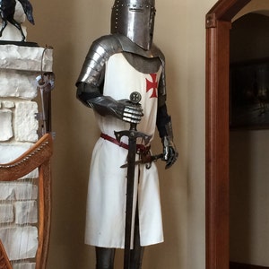 May include: A full-body suit of silver armor with a red cross on the chest. The suit is holding a sword and is standing on a wooden base.