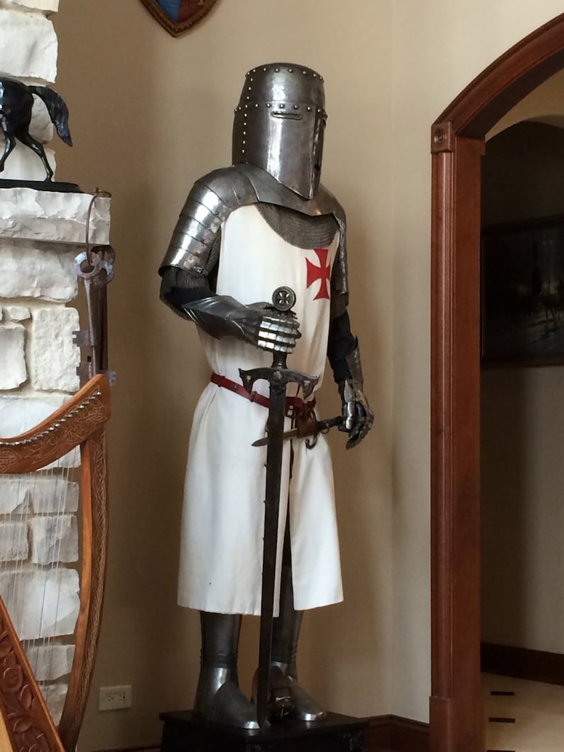 May include: A full-body suit of silver armor with a red cross on the chest. The suit is holding a sword and is standing on a wooden base.
