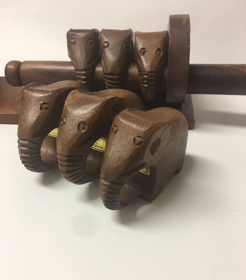 May include: Set of wooden elephant napkin rings with a wooden napkin holder. The napkin rings are brown and have a carved elephant design. The napkin holder is also brown and has a cylindrical shape.