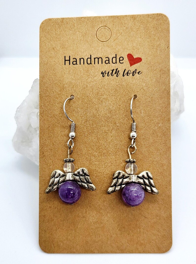 Puede incluir: Un par de pendientes de alas de &aacute;ngel hechos a mano con cuentas de piedras preciosas moradas. Los pendientes tienen alas y ganchos de metal plateado. La tarjeta dice "Handmade with love."