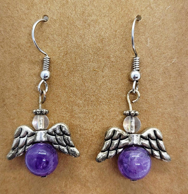 Puede incluir: Un par de pendientes colgantes con dijes de alas de &aacute;ngel y cuentas de piedras preciosas moradas. Los pendientes tienen ganchos y detalles plateados, con una cuenta transparente encima de las alas de &aacute;ngel. Las piedras preciosas son de color morado intenso.