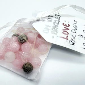 May include: A DIY bracelet kit containing pink rose quartz beads and two dark grey beads in a sheer white bag. A white tag attached to the bag reads "DIY KIT - BRACELET - LOVE: Rose Quartz". The kit is for making bracelets.
