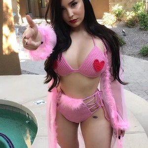 May include: A pink crochet bikini with a fuzzy trim and red heart accents. The bikini top has a halter style and the bottoms are high-waisted with a lace-up detail.