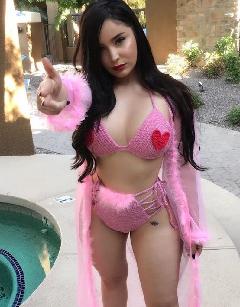 May include: A pink crochet bikini with a fuzzy trim and red heart accents. The bikini top has a halter style and the bottoms are high-waisted with a lace-up detail.
