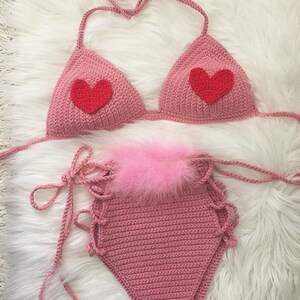 May include: A pink crocheted bikini top and bottom with red heart accents. The bikini top has adjustable straps and the bottom has a high-waisted style with a pink fluffy trim.