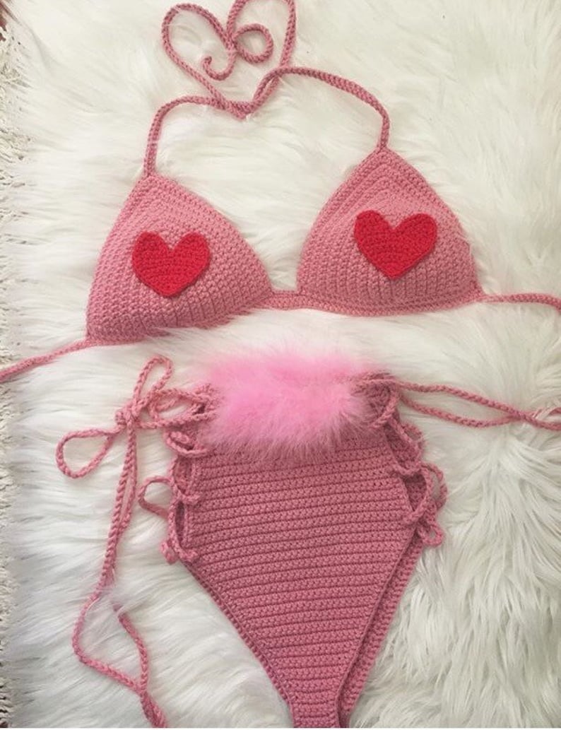 May include: A pink crocheted bikini top and bottom with red heart accents. The bikini top has adjustable straps and the bottom has a high-waisted style with a pink fluffy trim.
