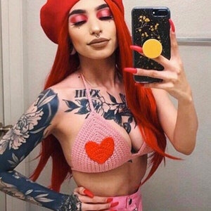 Puede incluir: Una mujer con cabello rojo y un gorro rojo está usando un top de bikini de crochet rosa con un corazón rojo en el centro. Tiene tatuajes en los brazos y sostiene un teléfono negro con un pop socket amarillo.