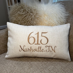 May include: A rectangular off-white linen throw pillow featuring brown embroidered text. The center displays the number 615 in a large serif font, with the text Nashville, TN embroidered in a smaller cursive script directly underneath it.