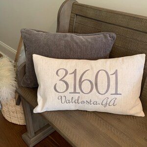 May include: A beige cushion with the text "31601 Valdosta, Ga" embroidered in grey on a wooden bench.