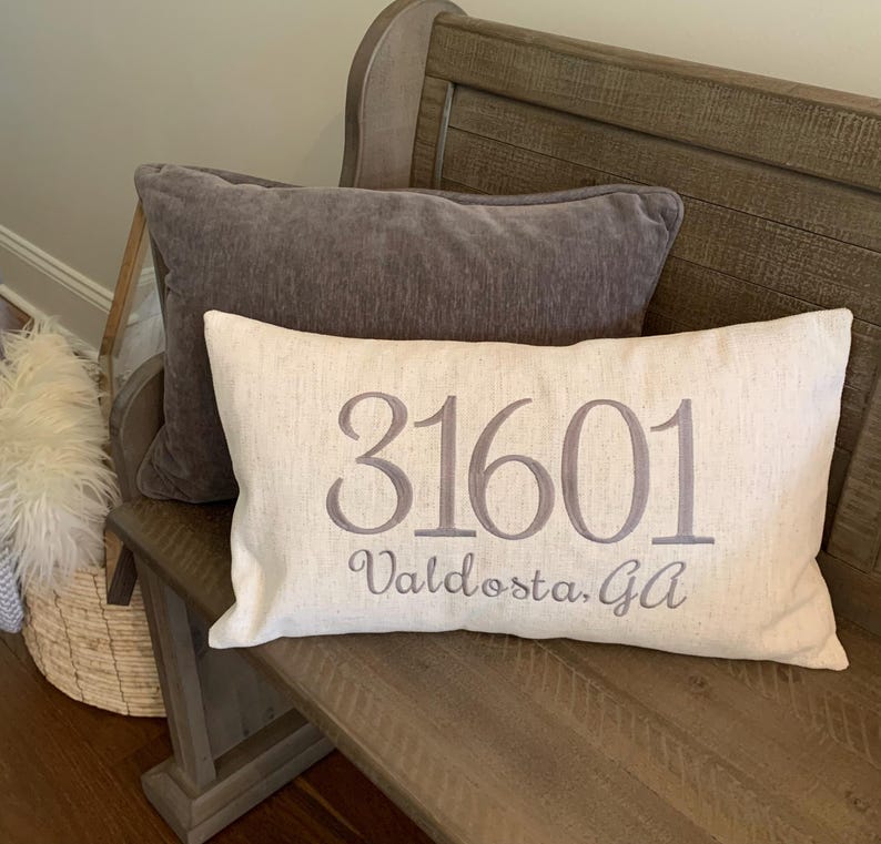May include: A beige cushion with the text "31601 Valdosta, Ga" embroidered in grey on a wooden bench.