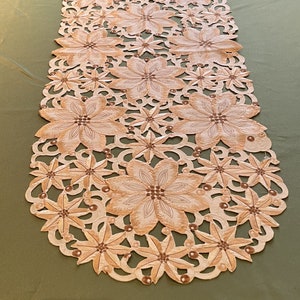 May include: A beige table runner with a delicate floral pattern and brown bead accents. The runner is oval shaped and has a lace-like design.