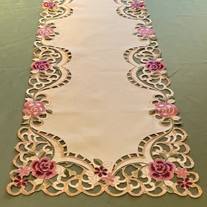 Puede incluir: Un camino de mesa beige con un delicado diseño floral. El camino de mesa presenta un patrón recortado con rosas bordadas en tonos de rosa y verde.