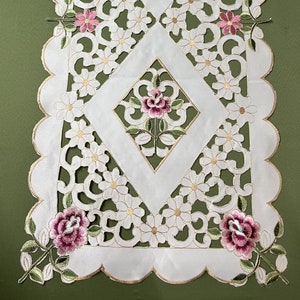 May include: A white rectangular table runner featuring intricate cutwork, scalloped edges, and embroidered floral motifs. The design includes repeating diamond shapes containing pink and purple rose blossoms with green leaves and small white flowers.