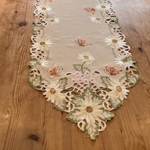 Puede incluir: Un camino de mesa beige con margaritas blancas bordadas, hojas verdes y mariposas naranjas. El camino de mesa tiene un borde festoneado con un delicado diseño de encaje.