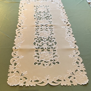 Può includere: Un runner da tavolo bianco con un delicato motivo di pizzo floreale. Il runner è realizzato in un tessuto morbido e leggero e presenta un bordo smerlato.