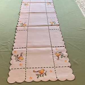 Peut inclure: Un chemin de table blanc avec un bord festonné et des oiseaux et des fleurs brodés. Le chemin de table est fait d'un tissu léger et a un design délicat.