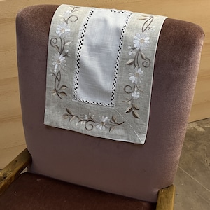 May include: A beige chair back with a white and beige embroidered chair back cover with a floral design. The chair is upholstered in a dark brown fabric.