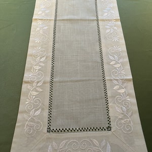 May include: Cream-coloured rectangular table runner with white embroidered floral and leaf designs. The centre has a plain, open weave fabric. The border features a decorative cut-out pattern. A decorative accent for a dining table, approximately 150 cm long.