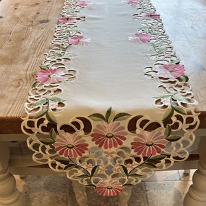 May include: A white table runner with a delicate floral design. The runner features pink and green embroidered flowers with a scalloped edge.