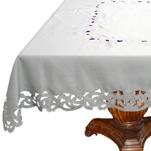 May include: White embroidered tablecloth with a delicate floral pattern around the edges. The tablecloth is draped over a wooden table with a pedestal base.