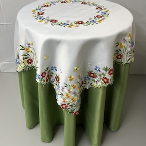 May include: White round tablecloth with embroidered floral design in shades of red, yellow, blue, and green. The tablecloth is draped over a green table.