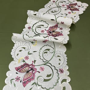 May include: White embroidered table runner with a scalloped edge. Features a repeating pattern of butterflies, flowers, and swirling vines in pink, yellow, and green. The runner is made of a delicate, sheer fabric.