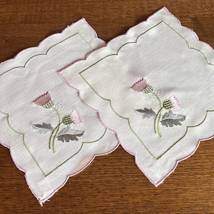 May include: Two white, square embroidered coasters with scalloped edges. Each coaster features a pink and green thistle design. The edges are trimmed in pink and outlined in green. The coasters are displayed on a wooden surface.