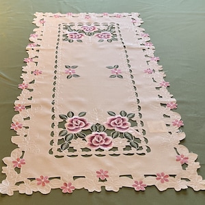 May include: White embroidered table runner with a delicate floral design featuring pink roses and leaves. The runner has a scalloped edge and a cut-out pattern.