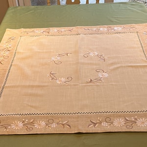 May include: A beige linen tablecloth with an embroidered floral design. The tablecloth has a scalloped edge and features a delicate floral pattern in shades of brown and white.
