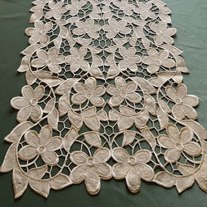 May include: White lace table runner with a delicate floral pattern. The runner is made of intricate lacework and features a repeating design of flowers and leaves.
