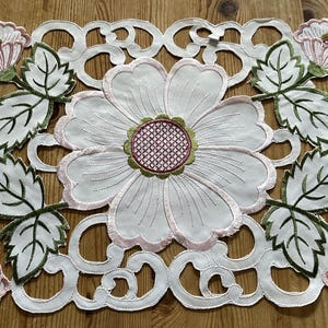 May include: White embroidered table runner with a floral design. The design features pink flowers with green leaves and a white background.