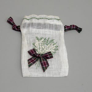 Puede incluir: Pequeño saco con cordón de color blanquecino con un diseño floral bordado en verde y un lazo a cuadros. El borde superior tiene costuras verdes y los cordones son de tela a cuadros a juego.