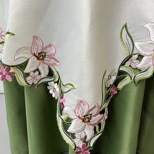 May include: White tablecloth with embroidered pink and green floral design. The tablecloth has a scalloped edge and is shown draped over a green tablecloth.