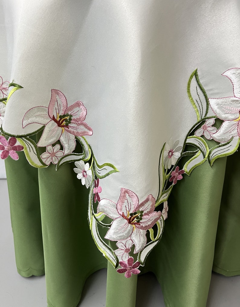 May include: White tablecloth with embroidered pink and green floral design. The tablecloth has a scalloped edge and is shown draped over a green tablecloth.