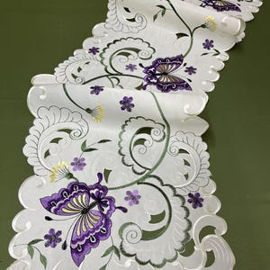 Può includere: Un runner da tavolo bianco con un ricamo di farfalle viola e viti verdi. Il runner ha un bordo a festoni.