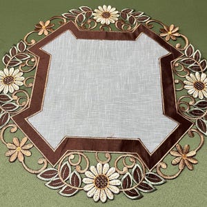 May include: Circular embroidered doily with a central geometric design and floral edging. The doily has a light brown border, a white centre, and decorative flowers and leaves in shades of brown and cream. The doily is placed on a green surface.